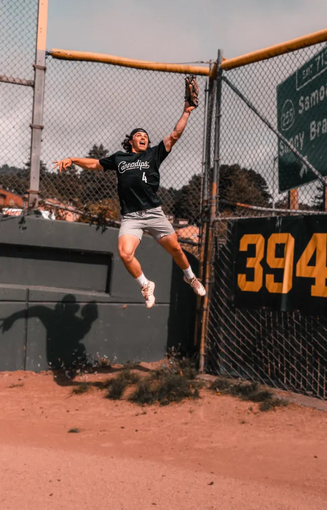 Player in a Cannadips shirt leaping for a catch at the outfield fence